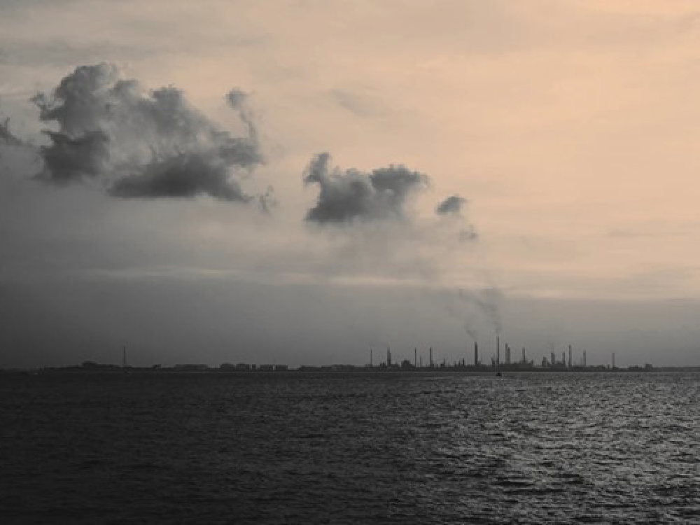 Dark sea with industrial skyline and smoke stacks under cloudy sky.